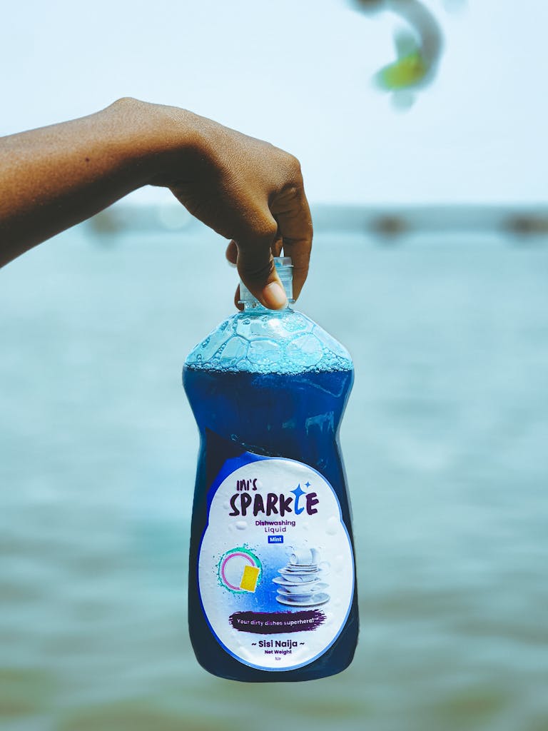 A hand holds a bottle of blue sparkling dishwashing liquid by the ocean, highlighting freshness.