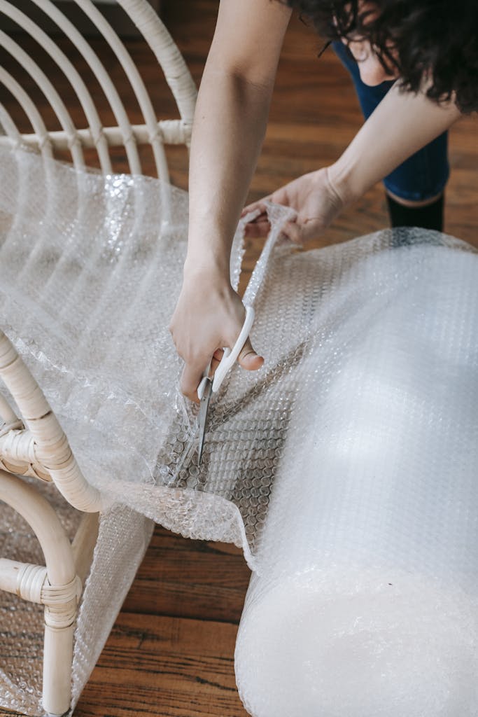 St. Augustine 9 A woman using scissors to cut bubble wrap for packing a wicker chair indoors, preparing to move.