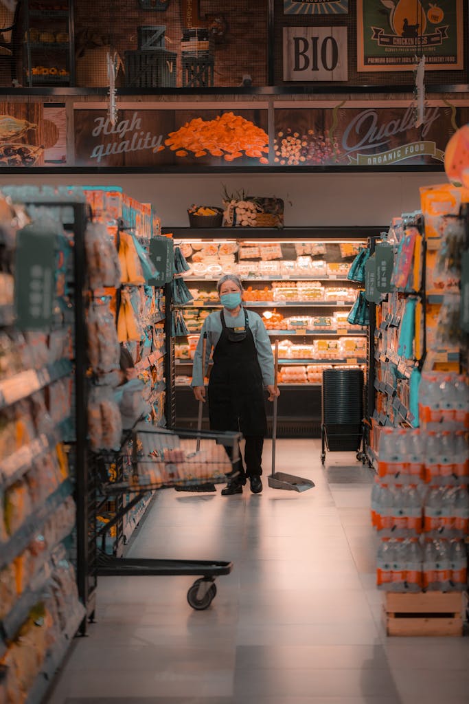 Elderly worker in mask cleans aisle of grocery store.