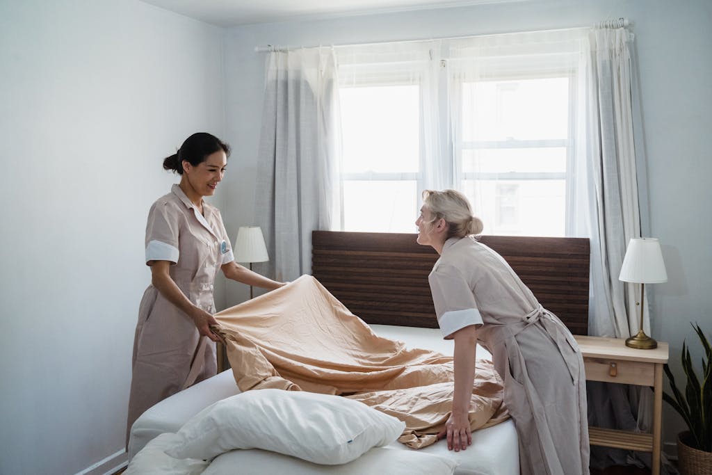 Two housekeepers in uniform make a bed in a well-lit bedroom, showcasing efficient housekeeping service.